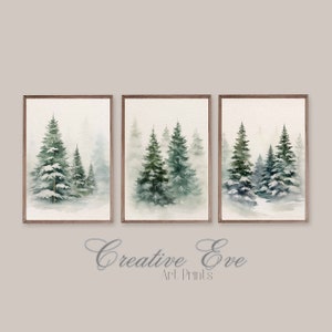 May include: Set of three framed watercolor art prints featuring winter forest scenes. Each print showcases evergreen trees in shades of green and blue, set against a misty, neutral background. The frames are a warm brown color. The text "Creative Eve Art Prints" is at the bottom.