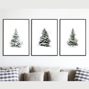 May include: Three framed prints featuring winter scenes. Each print showcases a snow-covered evergreen tree against a white background. The trees vary in size and shape, with a minimalist aesthetic. The frames are black, and the overall theme is winter decor.