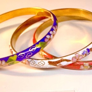 May include: Three gold-tone bangles with colorful enamel floral designs. One bangle is white with pink and purple flowers, another is red with flowers, and the third is blue with white accents.
