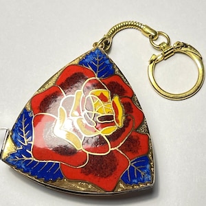 May include: A triangular, gold-toned keychain with a vibrant, enameled rose design. The rose features red, yellow, and white petals, set against a blue leaf background. The keychain includes a gold ring and a decorative, curved connector.