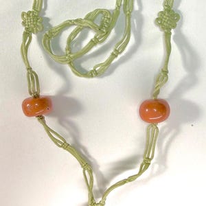 May include: A necklace featuring a light green braided cord with decorative knots, two smooth coral-colored beads, and a central pendant consisting of a smaller coral-colored bead above a carved, pale green stone figure with a smooth finish.