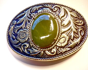 May include: Oval-shaped belt buckle with a central, olive-green stone. The buckle features intricate floral and scrollwork designs in silver and gold tones, with a dark blue border. The overall design is ornate and detailed.