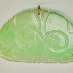 Puede incluir: Colgante de jade verde claro con diseños florales y geométricos tallados. El colgante tiene una anilla dorada en la parte superior para colgarlo. El jade tiene una calidad translúcida y un acabado pulido. La forma general es redondeada y estilizada.