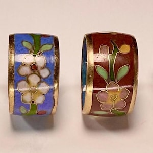 May include: Six decorative napkin rings with floral designs. Each ring features a different color scheme, including black, blue, red, and green, with gold trim and a turquoise interior. The rings are adorned with white and pink flower patterns.