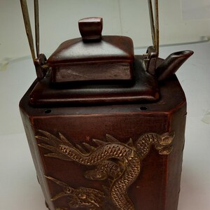 May include: A dark brown hexagonal teapot featuring a raised gold-toned dragon relief on its side. The teapot has a square lid with a central knob, a short spout, and a tall, thin metal swing handle attached to the top edges of the vessel.