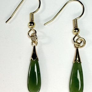 Puede incluir: Un par de pendientes colgantes dorados. Cada pendiente presenta una piedra de jade verde oscuro en forma de lágrima suspendida de un gancho dorado. Las piedras de jade tienen un acabado pulido y brillante, sobre un fondo blanco.