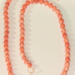 May include: A long strand of smooth, round, salmon-pink coral beads arranged in a continuous loop. The beads feature subtle variations in color and texture, with a light-colored string visible at the ends where the strand is tied together.
