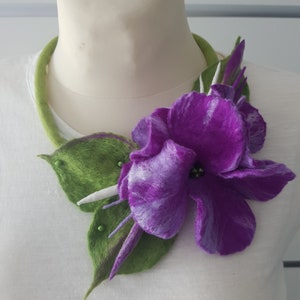 Puede incluir: Una flor de fieltro morado y blanco con hojas de fieltro verde, adornada con peque&ntilde;as cuentas blancas, est&aacute; unida a un collar de fieltro verde.