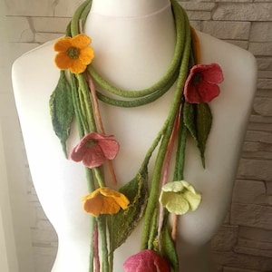 Puede incluir: Un collar de fieltro verde con flores de fieltro en tonos de amarillo, rosa y verde. El collar se muestra en un maniquí blanco.
