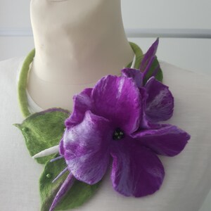 Puede incluir: Una flor de fieltro morado con hojas de fieltro verde, unida a un collar de fieltro verde. La flor tiene un centro blanco con cuentas negras.