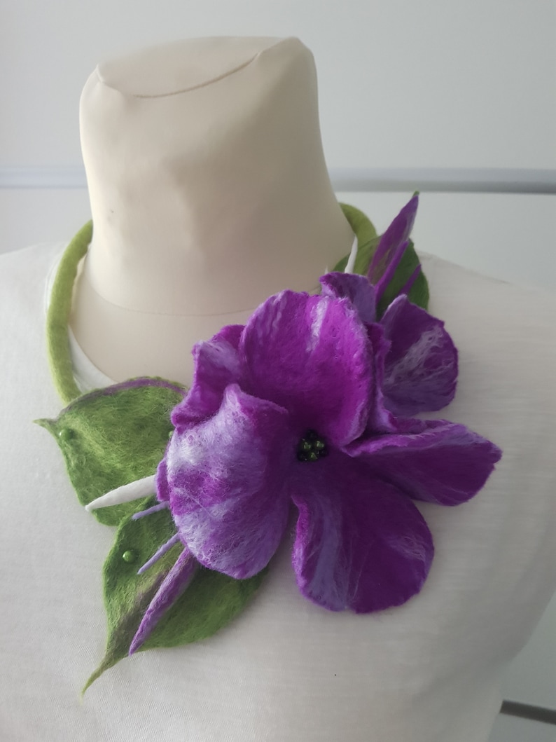 Puede incluir: Una flor de fieltro morado con hojas de fieltro verde, unida a un collar de fieltro verde. La flor tiene un centro blanco con cuentas negras.