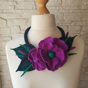 Puede incluir: Collar de fieltro con una gran flor morada y hojas verdes. La flor tiene un centro verde y es el punto focal del collar. El collar se exhibe en un maniquí blanco.