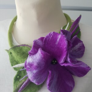 Puede incluir: Una flor de fieltro morado con hojas de fieltro verde, adornada con peque&ntilde;as cuentas verdes, est&aacute; unida a un collar de fieltro verde. El collar se muestra en un maniqu&iacute; blanco.