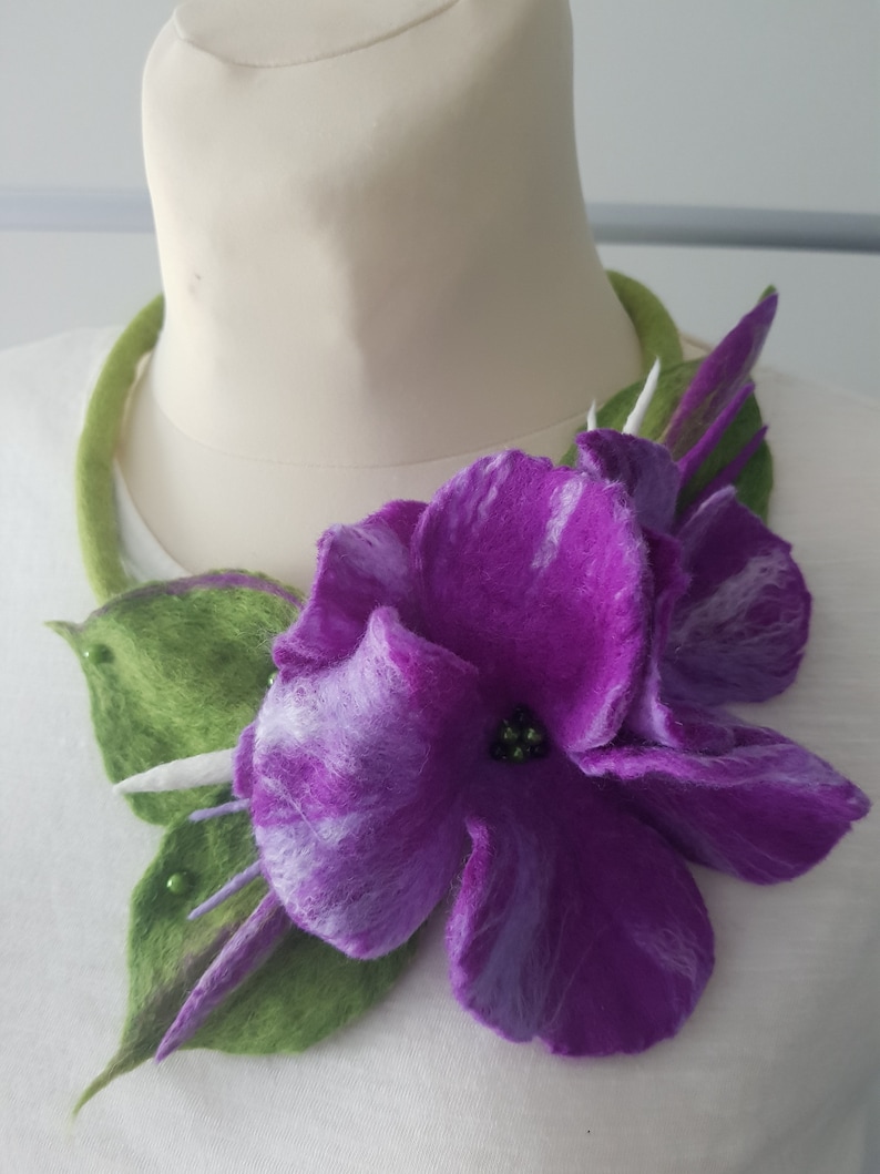Puede incluir: Una flor de fieltro morado con hojas de fieltro verde, adornada con peque&ntilde;as cuentas verdes, est&aacute; unida a un collar de fieltro verde. El collar se muestra en un maniqu&iacute; blanco.