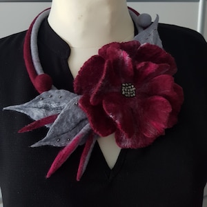 Puede incluir: Un collar de fieltro gris y burdeos con una flor de fieltro burdeos, hojas de fieltro gris y un centro de cuentas plateadas.