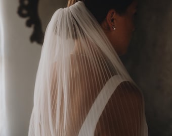 May include: A bride wearing a white pleated veil and a white dress. The veil is draped over the head and shoulders, creating a soft, flowing effect. The image is taken from a side profile, with a dark background.