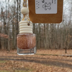 May include: A gold glitter car air freshener with a gingerbread man design. The air freshener is hanging from a brown tag that says "Sunshine Kisses Wax Studio LLC" and "Gingerbread". The tag also says "Hand Made in Ohio, Non-edible" and "sunshinekissesstudio.etsy.com".