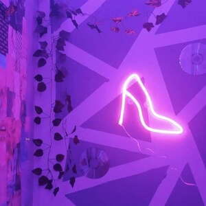 May include: A purple room with a neon pink high heel sign on the wall. The wall is decorated with a geometric pattern and a collage of images. There are also some green vines hanging from the wall.