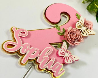 May include: Pink and gold cake topper with the name "Sophia" and a number "2" decorated with butterflies and flowers.