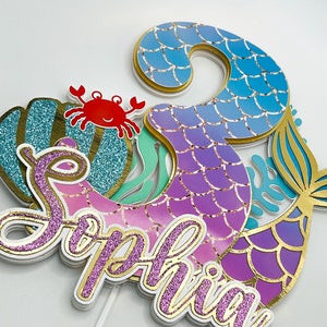 May include: A mermaid-themed cake topper with a pink, purple, and blue mermaid tail, a gold glitter seashell, a red crab, and the name "Sophia" in pink glitter letters.