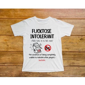 May include: White t-shirt with a humorous graphic design. The design features a milk carton with a face and the words "Fucktose Intolerant" with a phonetic pronunciation. The graphic also includes a red circle with a line through it and the letters "BS" inside.