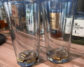 May include: Two clear glass tumblers filled with a clear liquid and several gold-coloured bullet casings. The glasses are on a wooden surface.