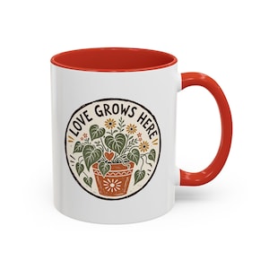May include: A white ceramic mug with a bright red interior and handle. The front features a circular graphic of a potted plant with green leaves, small yellow flowers, and a red heart, surrounded by the text LOVE GROWS HERE in black letters.