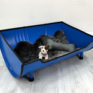 Op de afbeelding: Een blauw metalen hondenbed met een zwarte rand en vier poten. Het bed is gevuld met een grijs kunstbont bed en een pluche bot speelgoed.