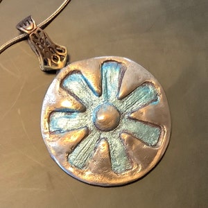 Peut inclure: Un collier avec un pendentif en argent et un charm rond de couleur cuivre repr&eacute;sentant une fleur stylis&eacute;e &agrave; huit p&eacute;tales. La fleur est recouverte d'un &eacute;mail bleu-vert et poss&egrave;de un centre circulaire sur&eacute;lev&eacute;.