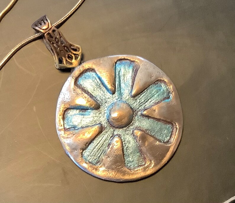 Peut inclure: Un collier avec un pendentif en argent et un charm rond de couleur cuivre repr&eacute;sentant une fleur stylis&eacute;e &agrave; huit p&eacute;tales. La fleur est recouverte d'un &eacute;mail bleu-vert et poss&egrave;de un centre circulaire sur&eacute;lev&eacute;.