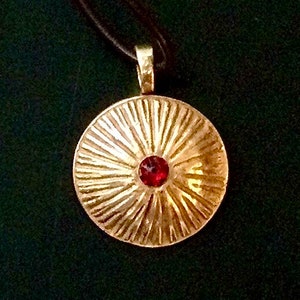 Könnte beinhalten: Goldfarbene Halskette mit einem runden Anhänger und einem roten Edelstein in der Mitte. Der Anhänger hat ein strukturiertes, sonnenstrahlenartiges Muster und hängt an einer schwarzen Kordel.