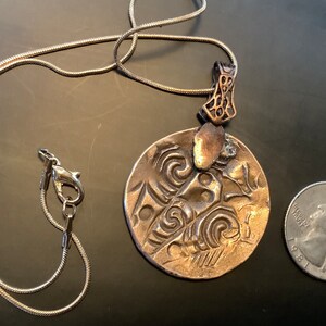 Peut inclure: Un collier en cha&icirc;ne argent&eacute;e avec un pendentif rond en bronze orn&eacute; d'un motif en relief complexe. Le pendentif est &agrave; peu pr&egrave;s de la taille d'une pi&egrave;ce de 25 cents am&eacute;ricaine.