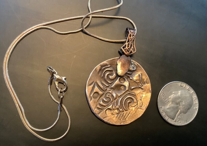 Peut inclure: Un collier en cha&icirc;ne argent&eacute;e avec un pendentif rond en bronze orn&eacute; d'un motif en relief complexe. Le pendentif est &agrave; peu pr&egrave;s de la taille d'une pi&egrave;ce de 25 cents am&eacute;ricaine.