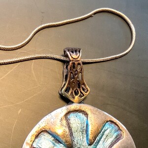 Peut inclure: Un collier en cha&icirc;ne argent avec un pendentif argent et or. Le pendentif est un disque rond avec un motif bleu et argent. Le pendentif a une b&eacute;li&egrave;re orn&eacute;e et d&eacute;taill&eacute;e.