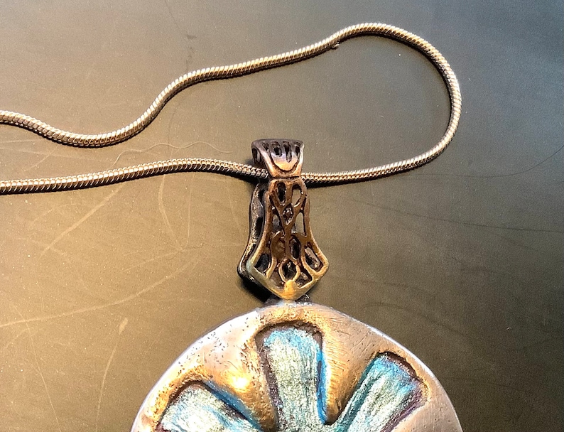 Peut inclure: Un collier en cha&icirc;ne argent avec un pendentif argent et or. Le pendentif est un disque rond avec un motif bleu et argent. Le pendentif a une b&eacute;li&egrave;re orn&eacute;e et d&eacute;taill&eacute;e.