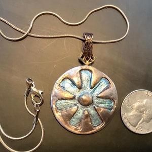 Peut inclure: Un collier en cha&icirc;ne argent avec un grand pendentif rond. Le pendentif est en cuivre et pr&eacute;sente un motif bleu et or. Le pendentif est &agrave; peu pr&egrave;s de la taille d'une pi&egrave;ce de 2 euros.