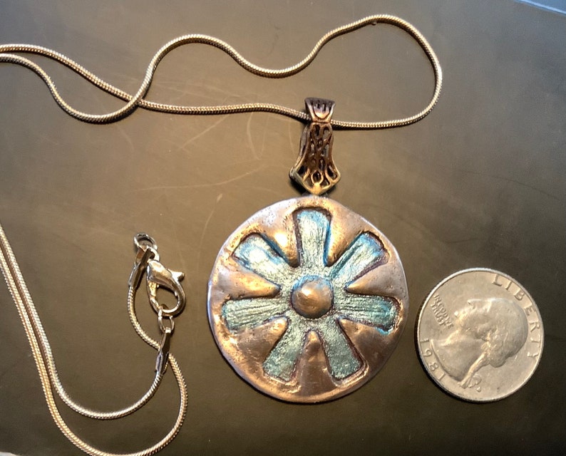 Peut inclure: Un collier en cha&icirc;ne argent avec un grand pendentif rond. Le pendentif est en cuivre et pr&eacute;sente un motif bleu et or. Le pendentif est &agrave; peu pr&egrave;s de la taille d'une pi&egrave;ce de 2 euros.