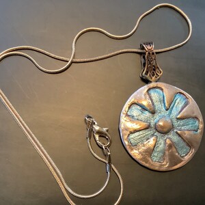 Peut inclure: Un collier en cha&icirc;ne argent avec un pendentif argent et bleu en forme de fleur. Le pendentif a un centre sur&eacute;lev&eacute; et six p&eacute;tales.
