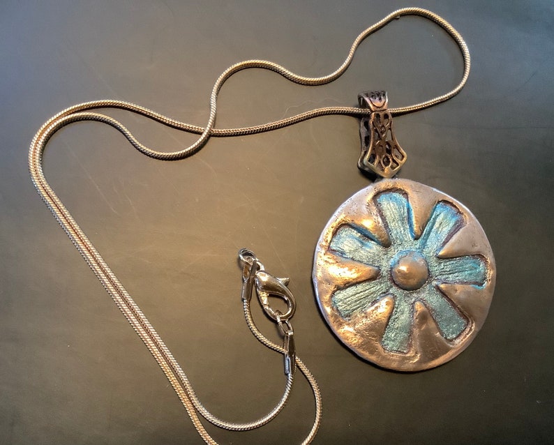 Peut inclure: Un collier en cha&icirc;ne argent avec un pendentif argent et bleu en forme de fleur. Le pendentif a un centre sur&eacute;lev&eacute; et six p&eacute;tales.
