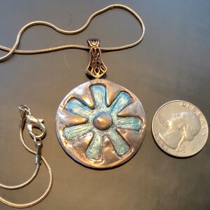 Peut inclure: Un collier avec un pendentif rond en argent, orn&eacute; d'une fleur bleue et argent&eacute;e. Le pendentif est &agrave; peu pr&egrave;s de la taille d'une pi&egrave;ce de 25 centimes d'euro.