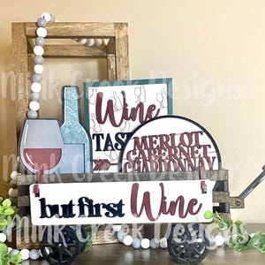 May include: A wooden wagon with a sign that reads "but first wine" in black and red lettering. The wagon is decorated with wine-themed decorations, including a wine bottle, a wine glass, and a round sign with the words "Merlot, Cabernet, Chardonnay" in red lettering.