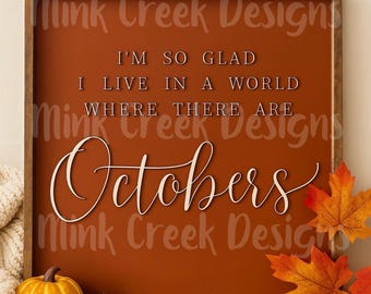 May include: A framed sign with the text "I'm so glad I live in a world where there are Octobers." The sign is brown with orange text and a brown wooden frame. Decorative pumpkins, pinecones, and fall leaves are in the foreground.