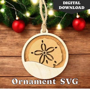 May include: A wooden Christmas ornament shaped like a sand dollar with a cutout design. The ornament is hanging from a twine string and has a white background.