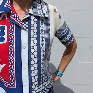Op de afbeelding: Een shirt met korte mouwen met een opvallend colorblock-ontwerp. De linkerkant heeft een rood, wit en blauw patroon, terwijl de rechterkant een blauw-witte geometrische print heeft. Het shirt heeft een klassieke kraag en witte knopen.