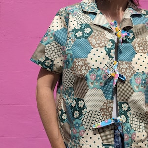 May include: A short-sleeved shirt with a patchwork design featuring various floral patterns in shades of blue, green, brown, and white. The shirt has a collar and is tied at the front with a ribbon.