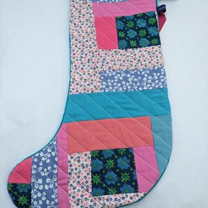 May include: A patchwork Christmas stocking with a blue, pink and green colour scheme. The stocking is quilted and has a small loop at the top.