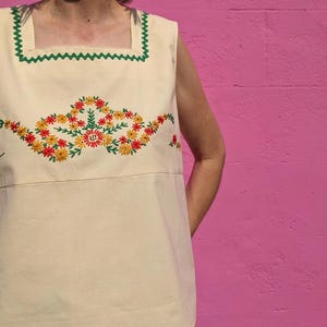 May include: A sleeveless, cream-colored top with a square neckline and green trim. The top features a floral embroidery design in shades of red, yellow, and green.