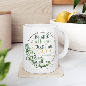 May include: White ceramic mug with a handle, featuring the text "Be still and know that I am GOD." in a circular design with green leaves and white flowers. The text "Psalm 46:10" is also included.