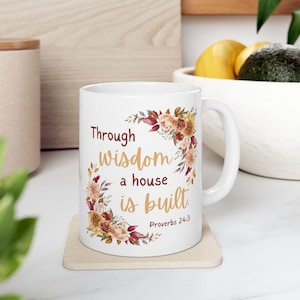 Pode incluir: Caneca de cerâmica branca com um design de coroa floral e o texto "Through wisdom a house is built" de Provérbios 24:3. A coroa apresenta flores em tons de pêssego, castanho e bordô. A caneca está sobre um descanso de copo bege.