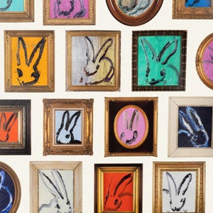 May include: A series of framed rabbit paintings in diverse colours, such as pink, blue, and orange. The artwork has a minimalist style with bold brushstrokes and gold frames. The paintings are set against a cream-coloured background.